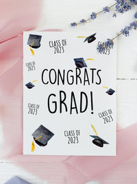 class of 2023 congrats grad graduation card. Graduation hats and class of 2023 arranged all over the face of the card. Black caps and gold tassels.